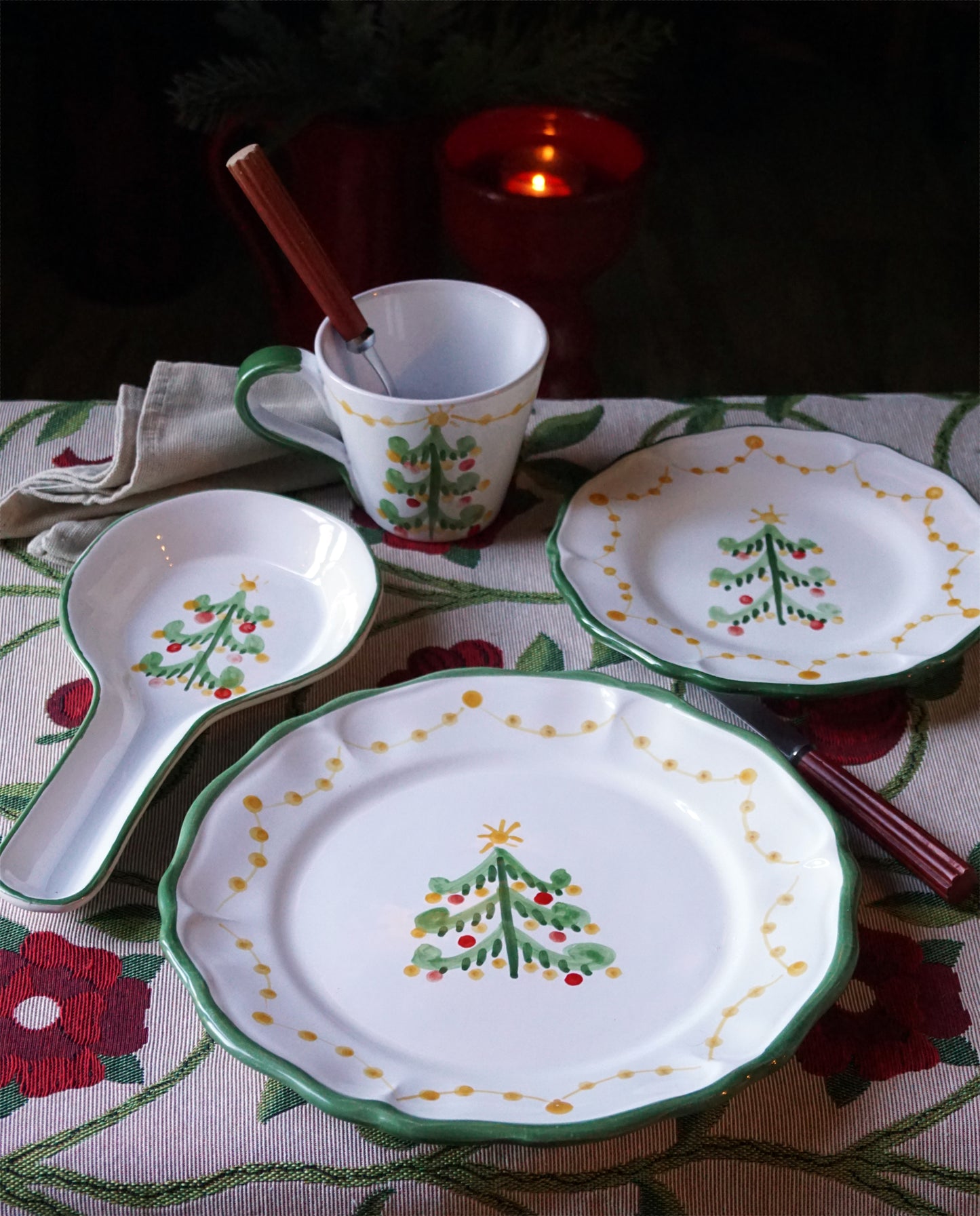 Set of ceramic plates, mug, and spoon rest with a green Christmas tree design on a patterned tablecloth.