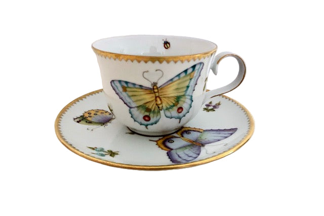 A porcelain tea cup with a butterfly and meadow pattern, including a gold rim and handle, displayed on a matching saucer.