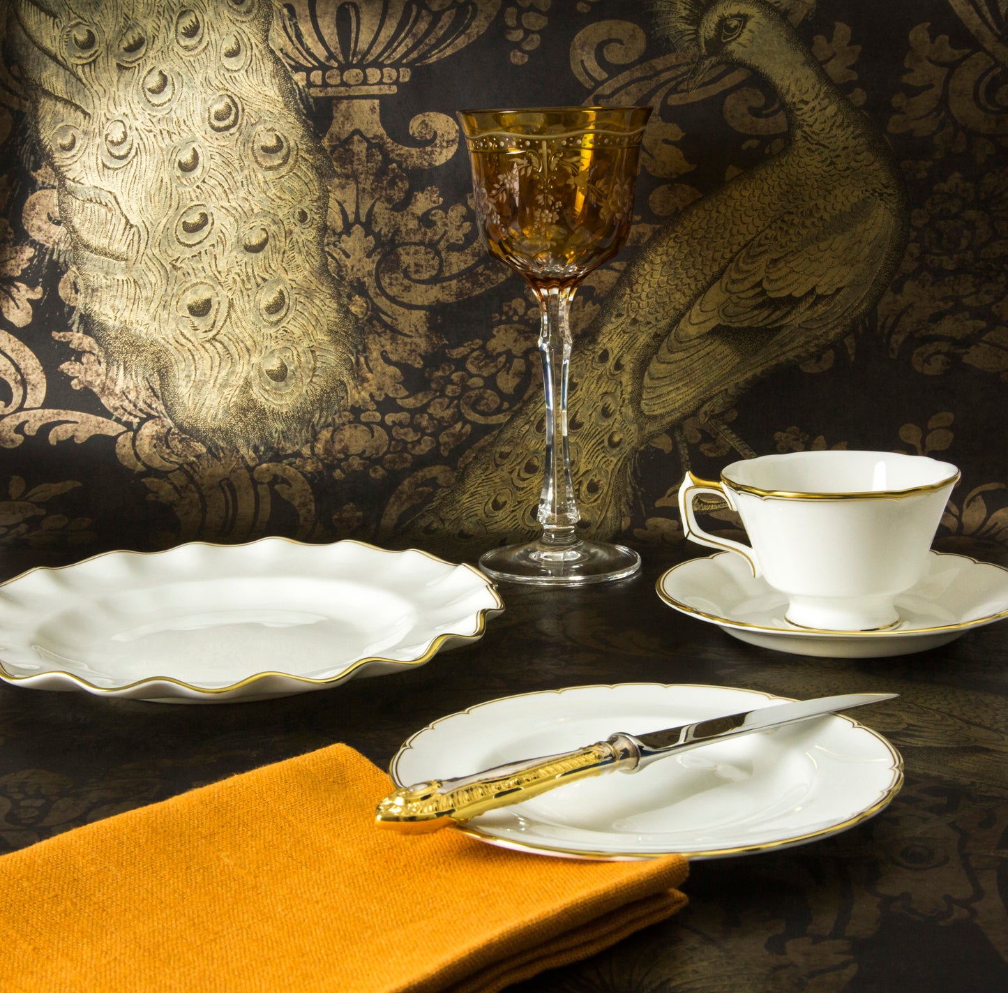 A set of fine bone china dinnerware featuring a classic white color with a luxurious scalloped gold rim, displayed on a table with a peacock feather patterned backdrop.