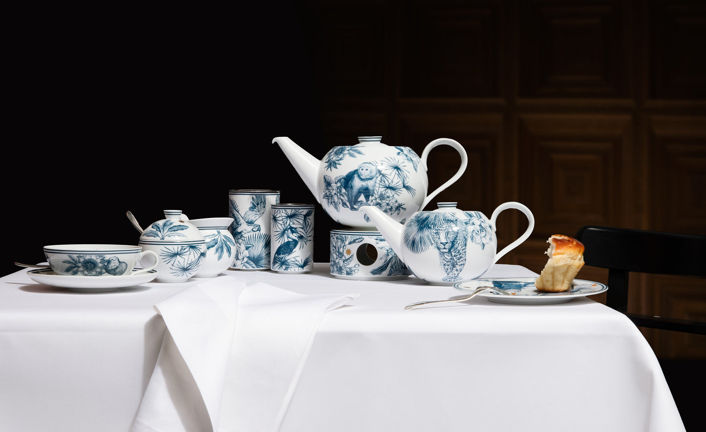 Tropical blue and white designed tea set with teapot, cups, and saucers on a white tablecloth against a dark background