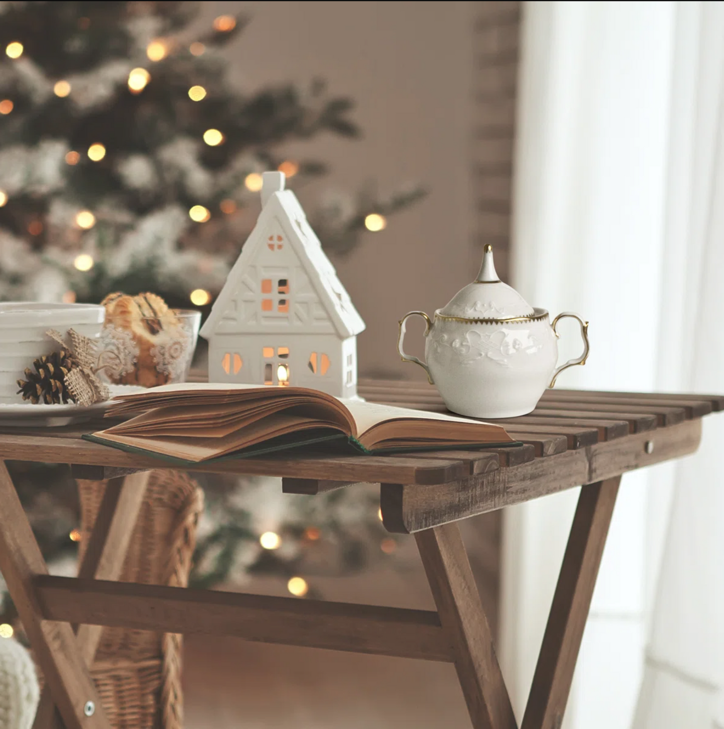 Decorative scene with a house-shaped candle, sugar bowl, and open book on a wooden table with a blurred Christmas tree in the background.
