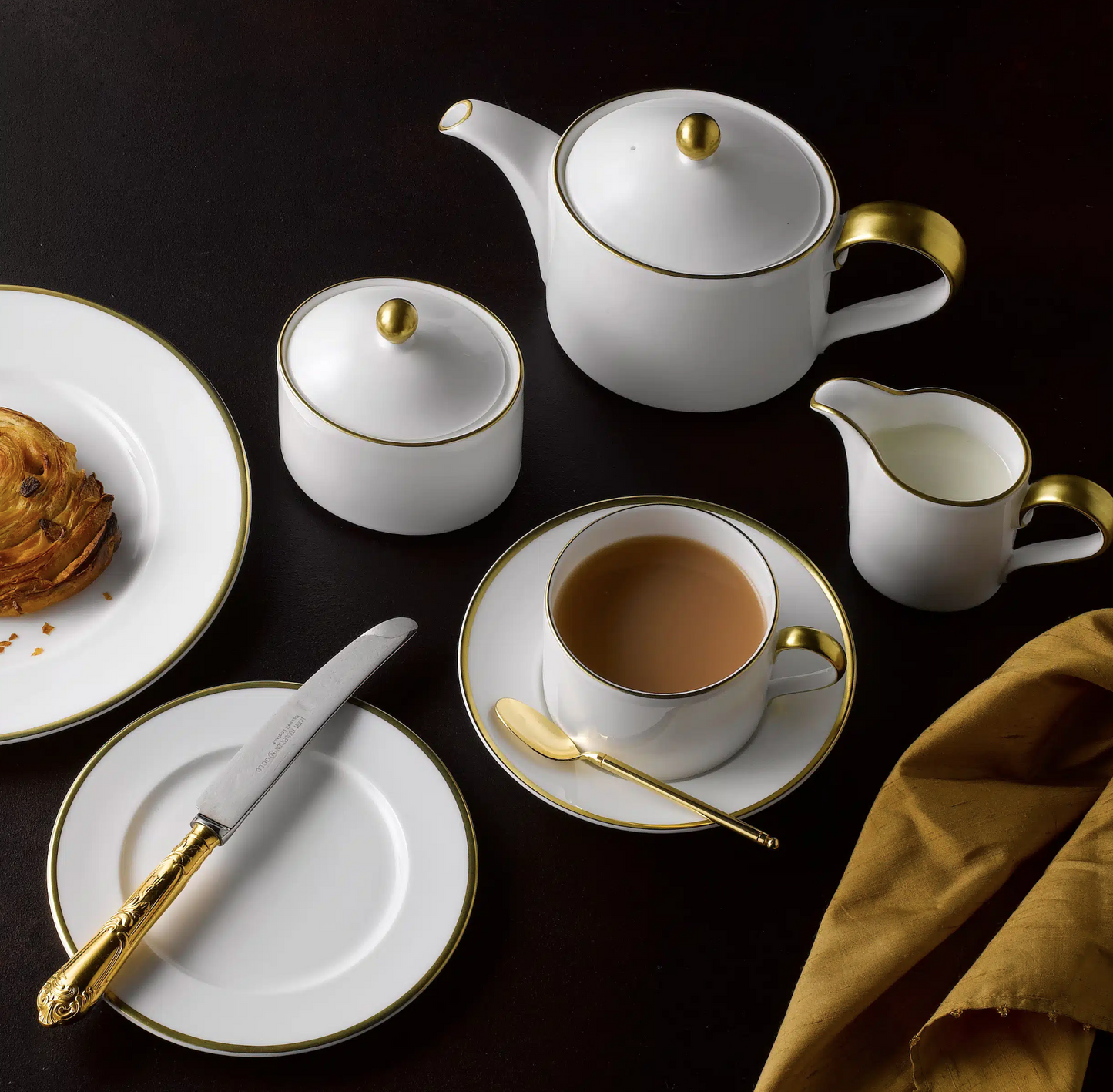 A set of white fine bone china drinkware with a 22 carat gold rim, including teacups, plates, and a teapot, displayed on a dark surface with a golden napkin on the side.
