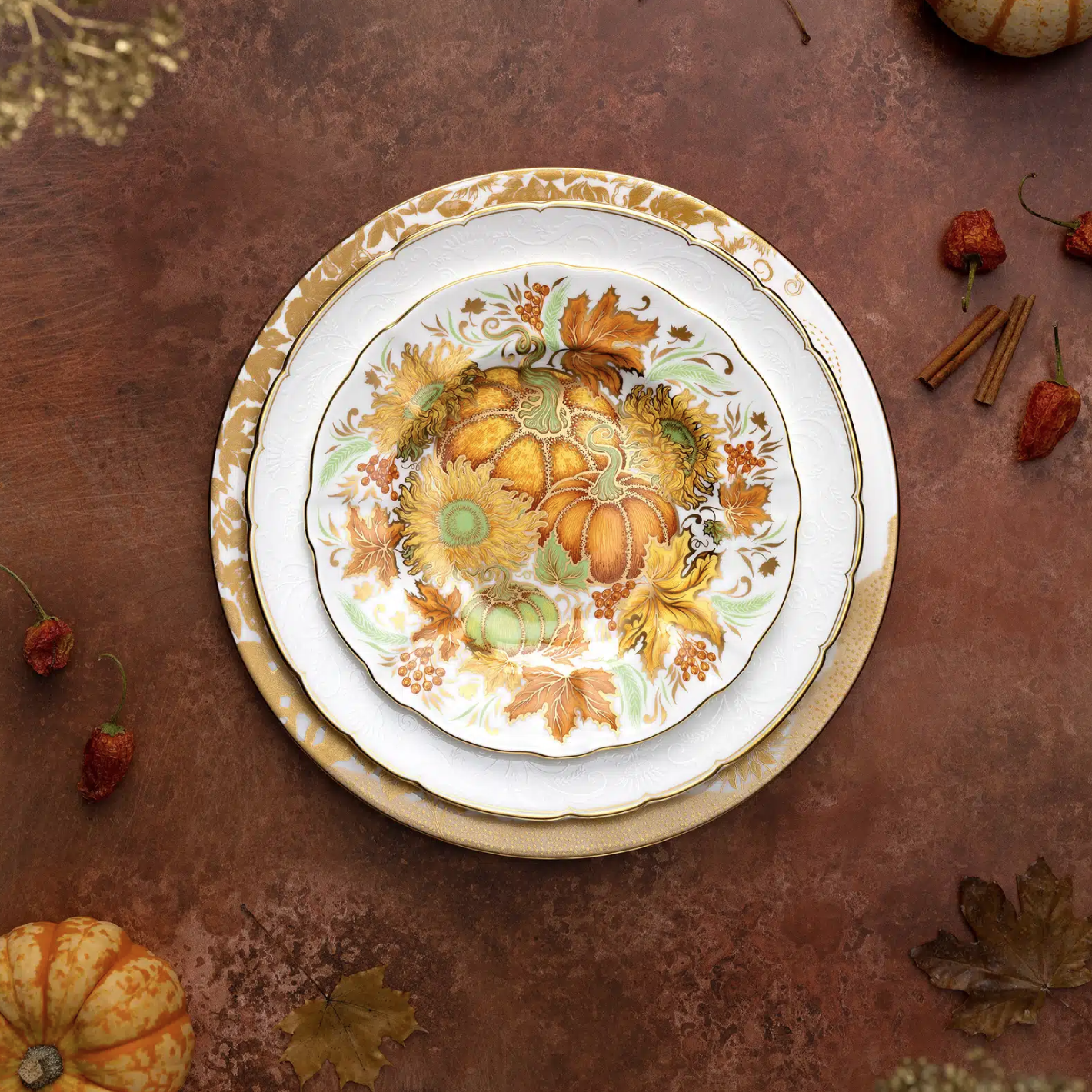 A set of fine bone china holiday accent plates with a multicolored floral and pumpkin design and gold accents, displayed on a table with autumn leaves and small pumpkins around them.