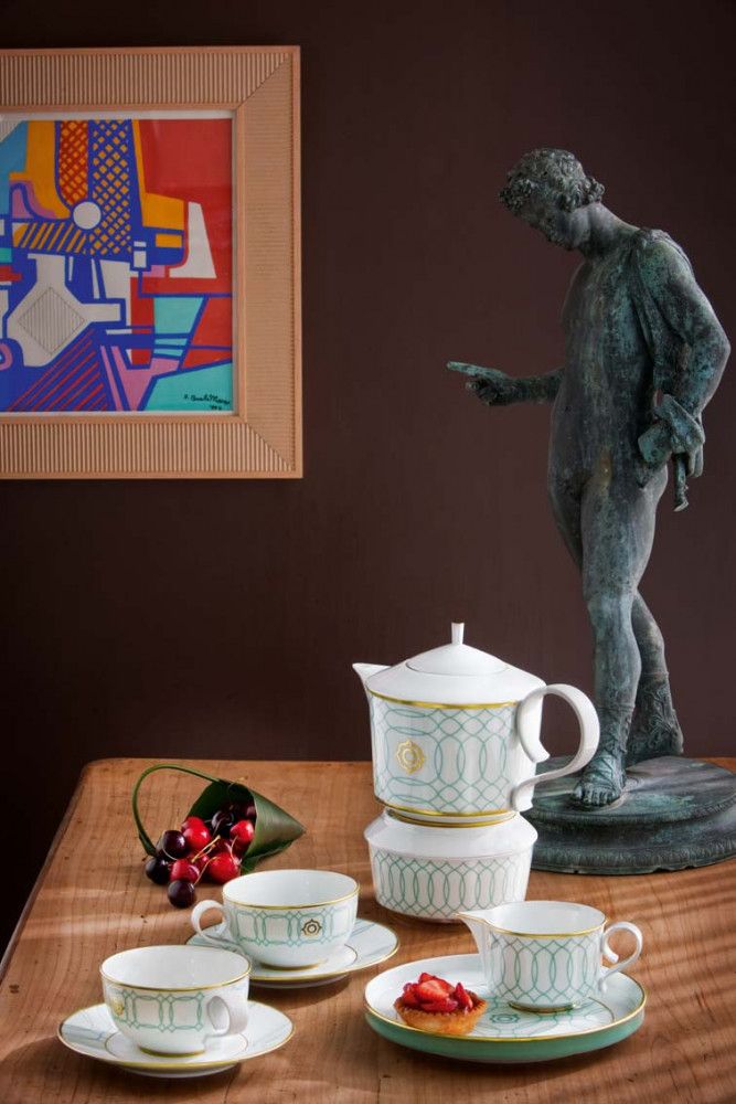 Tea set with teapot, cups, and saucers on a table with a statue and abstract painting in the background.