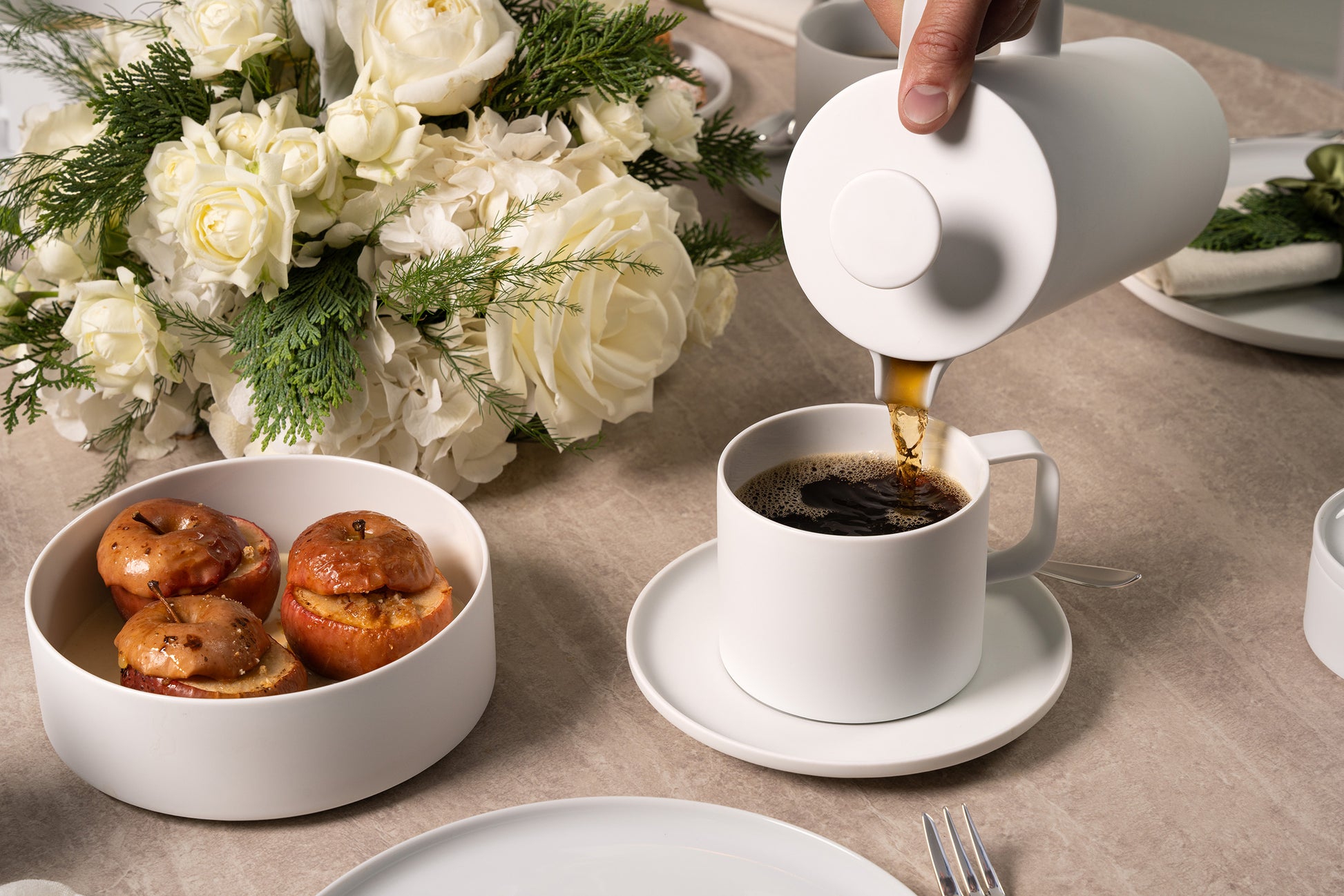 Person pouring coffee into a white cup on a table with flowers and pastries.