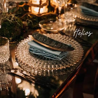 A styled table setting with a clear textured glass charger plate holding a folded slate blue napkin and a dark printed menu card, on a reflective dark table with warm candlelight and glassware in the background.