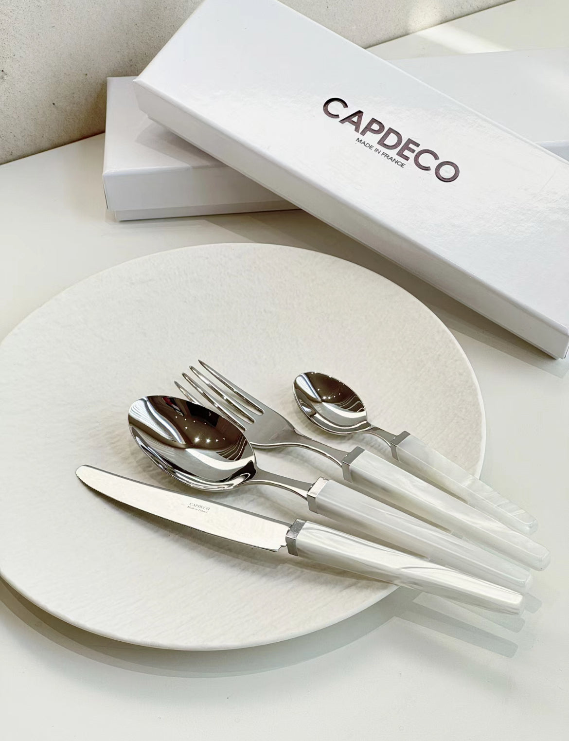 Silver cutlery set on a white plate with a 'Capdeo' branded box in the background.