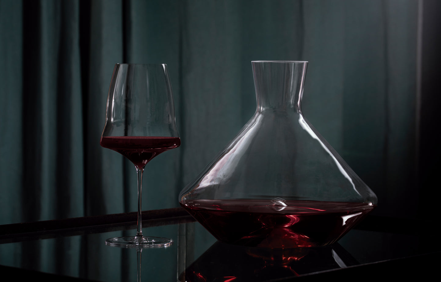 A clear glass decanter filled with red wine sits beside a matching hand-blown wine glass on a dark reflective surface against a dark teal curtain background.
