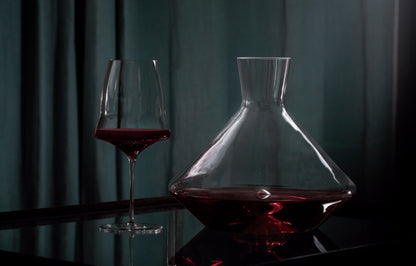 A clear glass decanter filled with red wine sits beside a matching hand-blown wine glass on a dark reflective surface against a dark teal curtain background.
