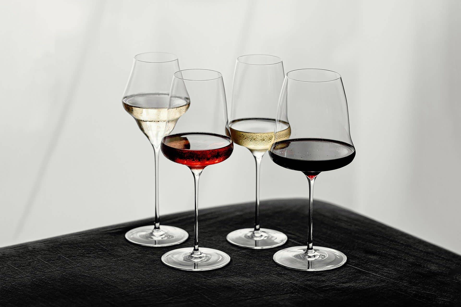 Four stemmed luxury wine glasses arranged on a dark surface against a light gray background, with white, red, and red wine partially filled in the glasses.