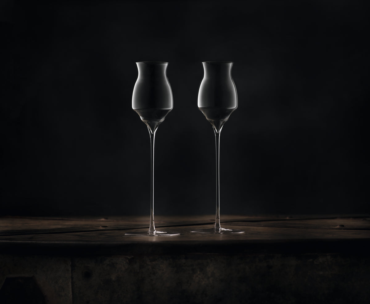 Two clear hand-blown, long-stemmed glasses for spirits with a distinctive flared, hourglass-shaped bowl and long stems, standing side-by-side on a dark rustic wooden surface against a dark background.