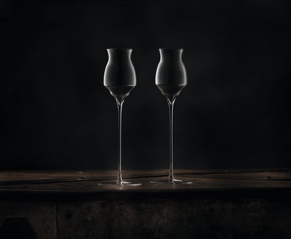 Two clear hand-blown, long-stemmed glasses for spirits with a distinctive flared, hourglass-shaped bowl and long stems, standing side-by-side on a dark rustic wooden surface against a dark background.