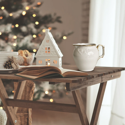 Cozy indoor setting with a wooden table, book, mug, and small house-shaped lantern with a Christmas tree in the background.