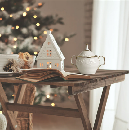 Decorative scene with a house-shaped candle, sugar bowl, and open book on a wooden table with a blurred Christmas tree in the background.