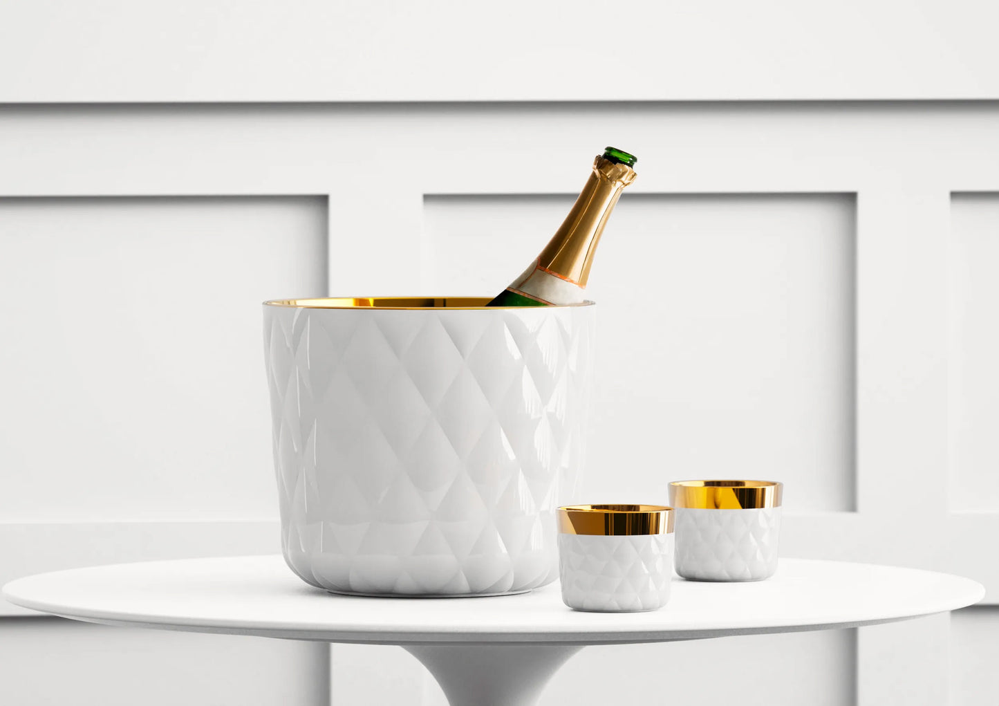 Bottle of champagne in a luxury white quilted texture ice bucket with matching cups on a white surface.