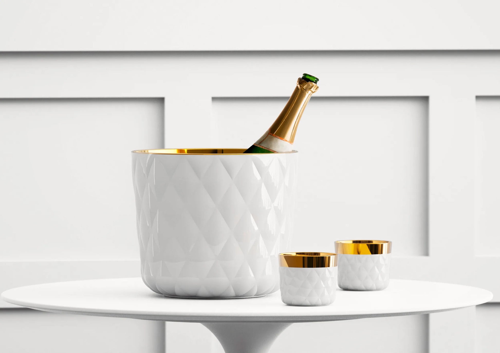Bottle of champagne in a luxury white quilted texture ice bucket with matching cups on a white surface.
