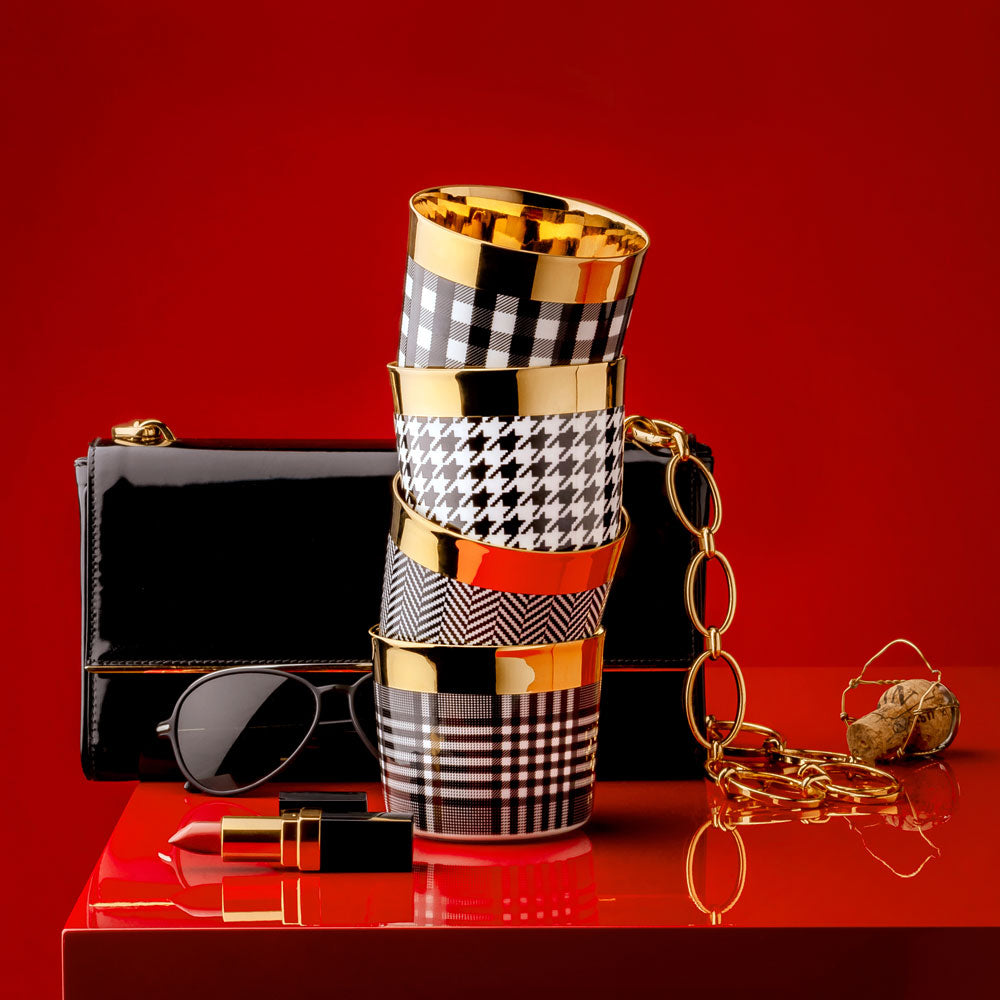 Luxury champagne tumblers with black and white designs and gold interiors stacked next to an elegant handbag, sunglasses, lipstick, and champagne bottle cork