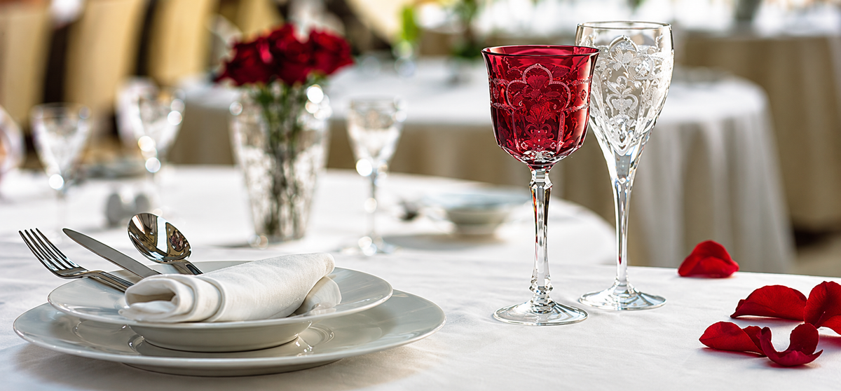 A styled dining table setting with white plates, a folded white napkin, silver cutlery, a red glass goblet, and a clear wine glass, with red rose petals on a white tablecloth.