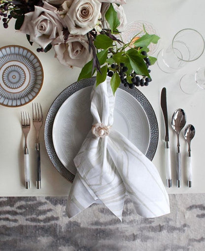 A styled table setting featuring a white patterned dinner plate with a textured gray charger plate, a white napkin tied with a small pearl, silver flatware, a floral centerpiece with pale roses, and clear drinking glasses on a light neutral background.