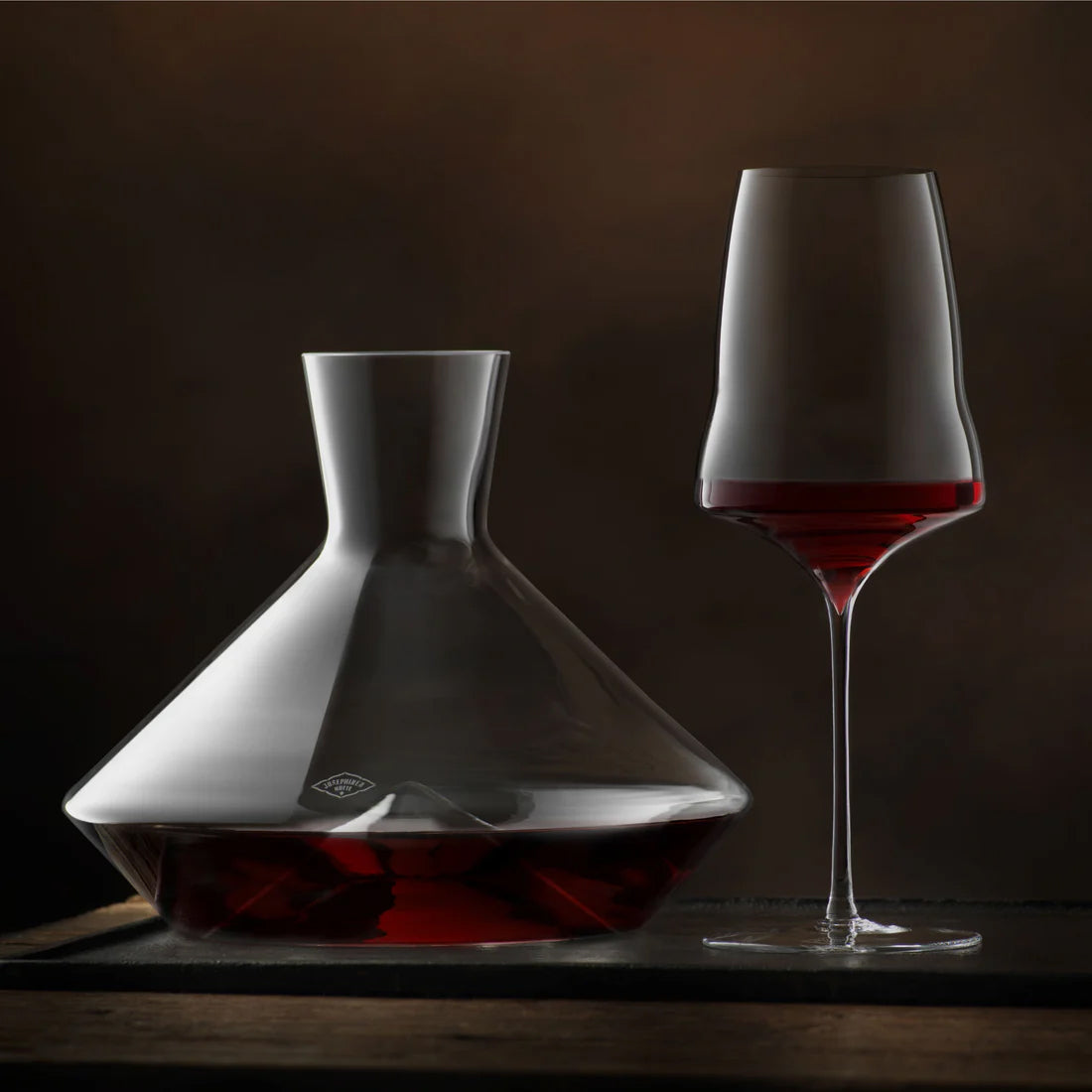 A clear glass decanter and matching wine glass on a dark wooden surface against a dark brown background. The decanter and glass are partially filled with red wine, which is visible in the glass and at the bottom of the decanter.